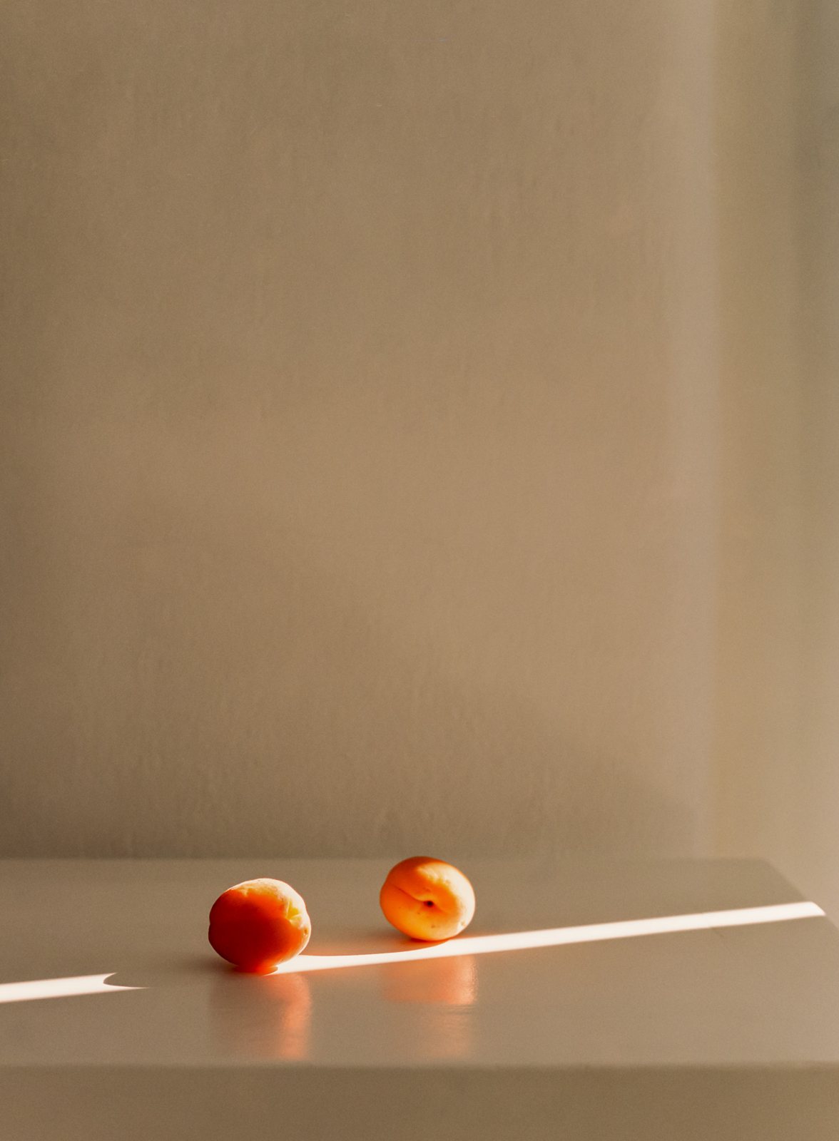 Two apricots rest on a pale table in a thin line of sunlight.