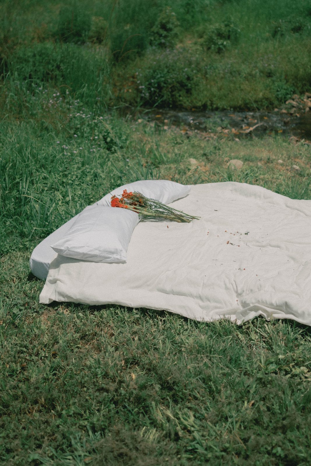 A white mattress and pillows lie on green grass beside a stream, with a bunch of orange flowers resting on the pillows.