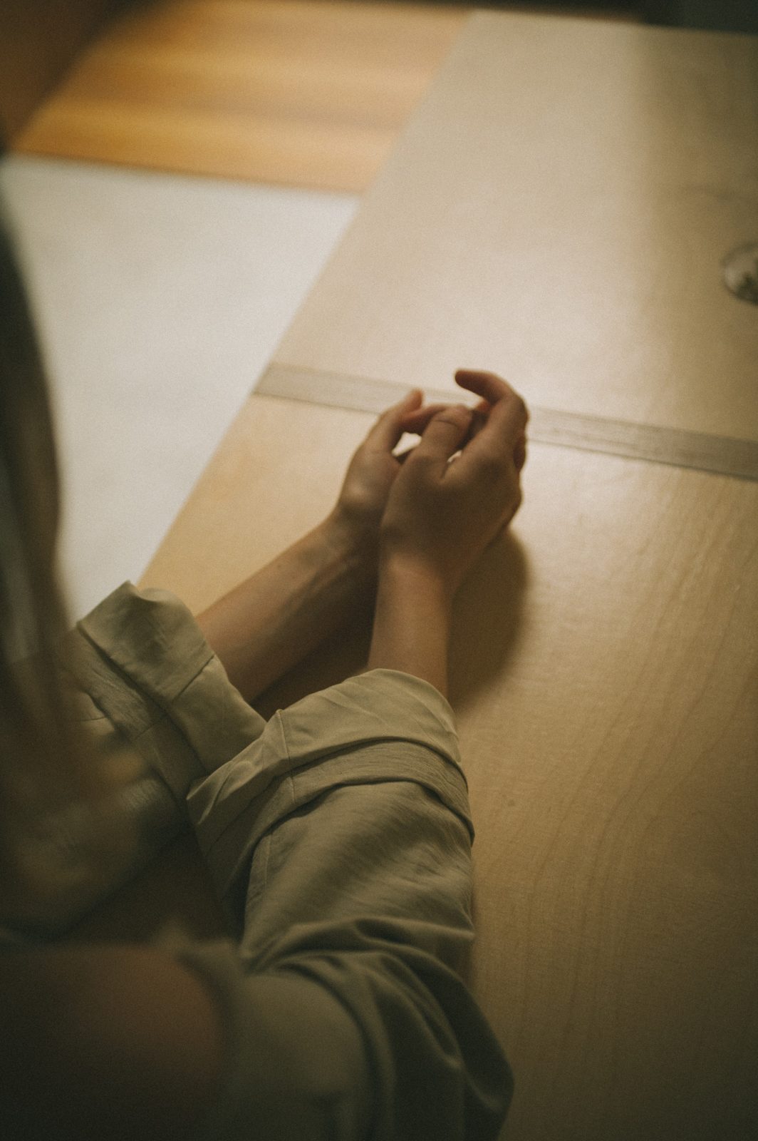A pair of hands rests loosely clasped on a pale wooden table.