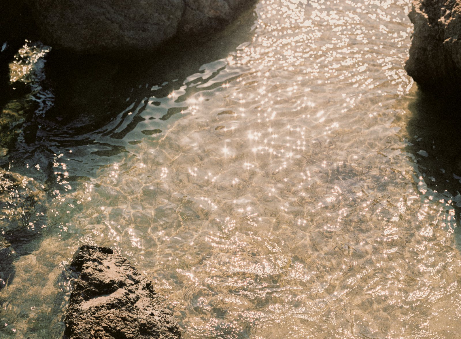 Sunlight glitters on clear, shallow water running between dark rocks.